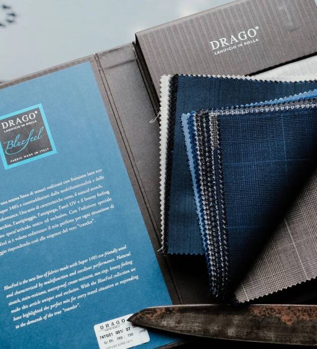 Vzorové látky Drago Bluefeel vystavené na desce. Různé odstíny modré a šedé látky z kolekce Super 140's eco-friendly wool, s informační kartou výrobce.